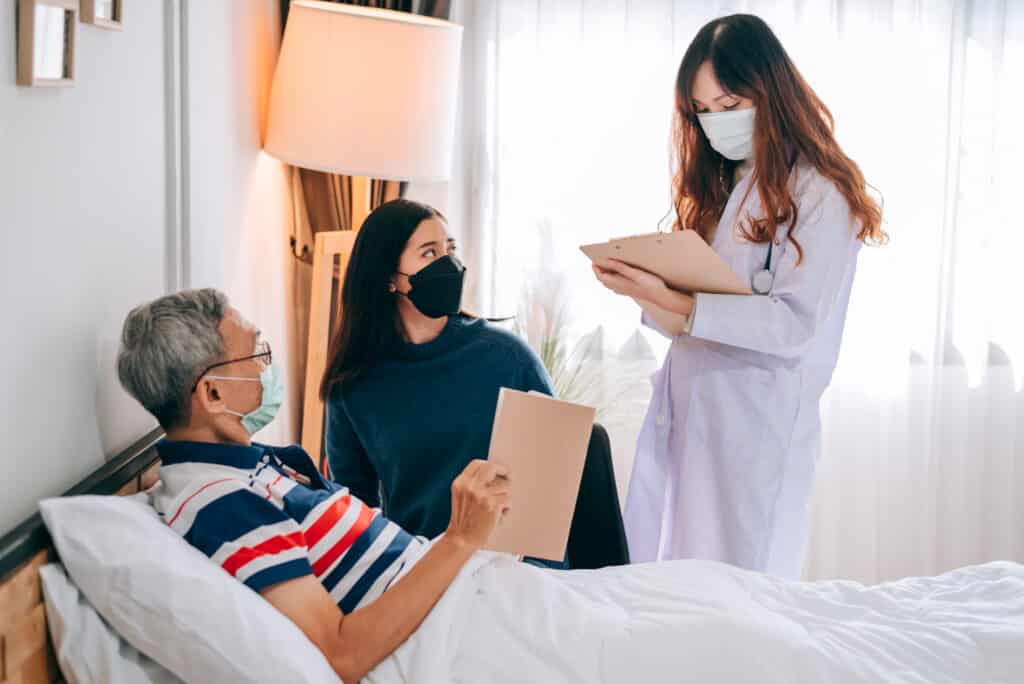 Asian doctor visit and examines a senior man at home. The grand-daughter is sitting next to the man. The three people are wearing masks to protect themselves.