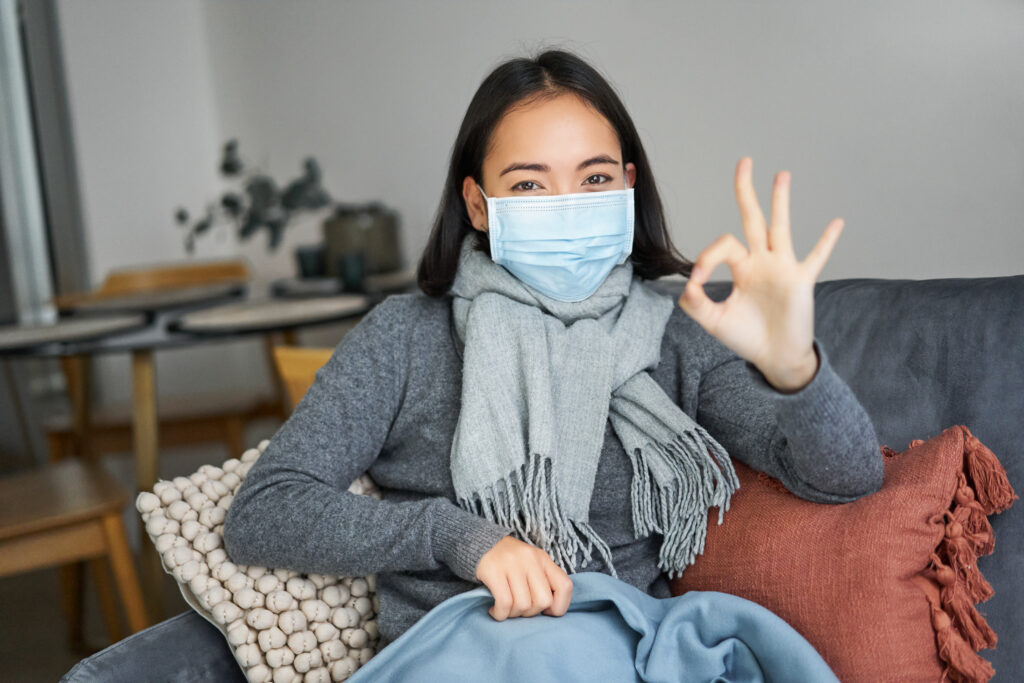 A young Asian woman wearing a mask gestures with her hand that all is well.