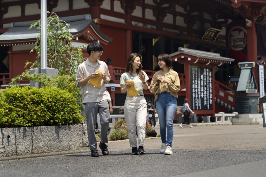 Three Asian friends go for a walk in Japan, one boy and two girls. They are walking down a street with a red temple.