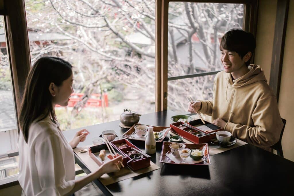 A Japanese man and woman sharing a meal together in a restaurant. They are smiling and enjoying the moment. They are seated near a window that looks out onto a cherry blossom tree.