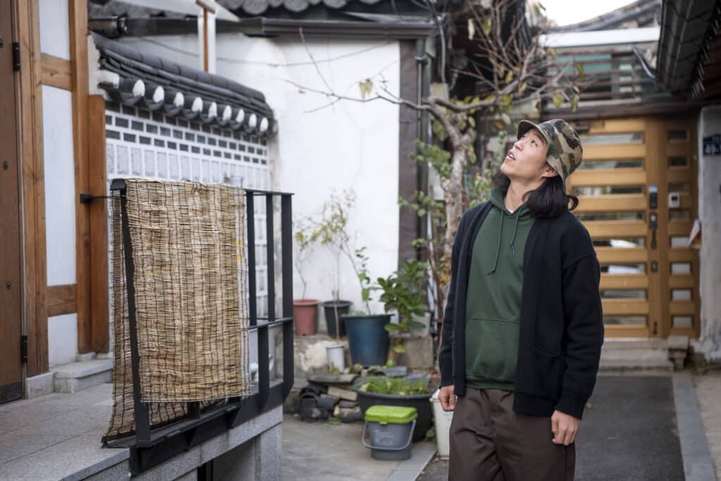 Japanese man in front of the entrance to a Japanese house, looking up to the first floor. He is wearing a hat and a black jacket, and seems interested in the building.