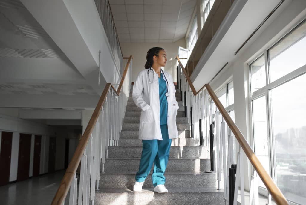 Black woman at work. She is wearing a doctor's smock and standing on the stairs of a health establishment.