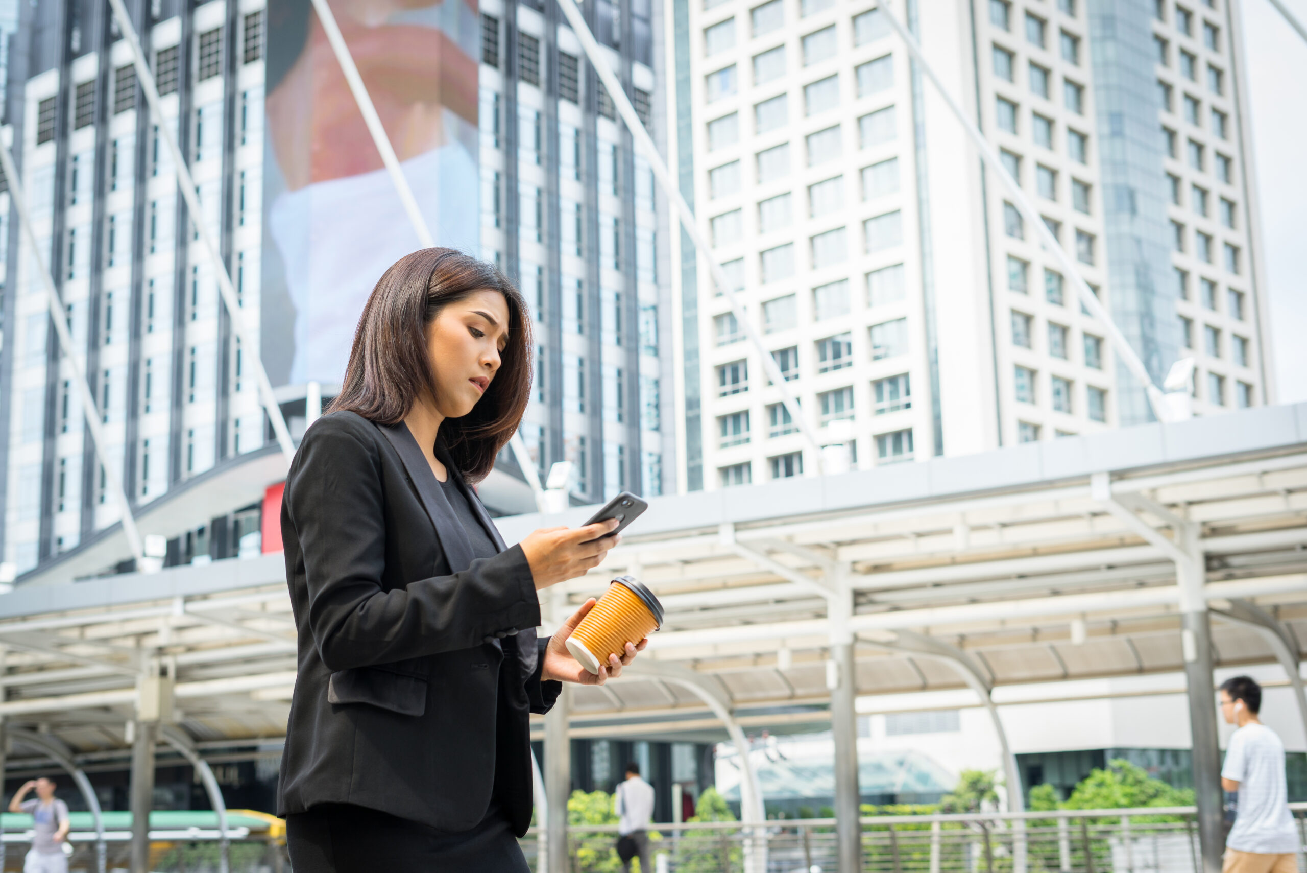 Businesswoman using a telephone with coffee in hand, walking down the street with office buildings in the background.