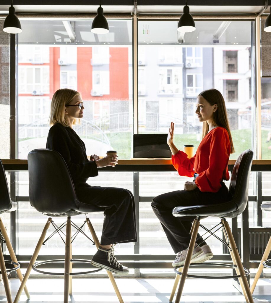 Two businesswomen have a conversation at a restaurant table over a cup of coffee. One woman gives the other a friendly wave.