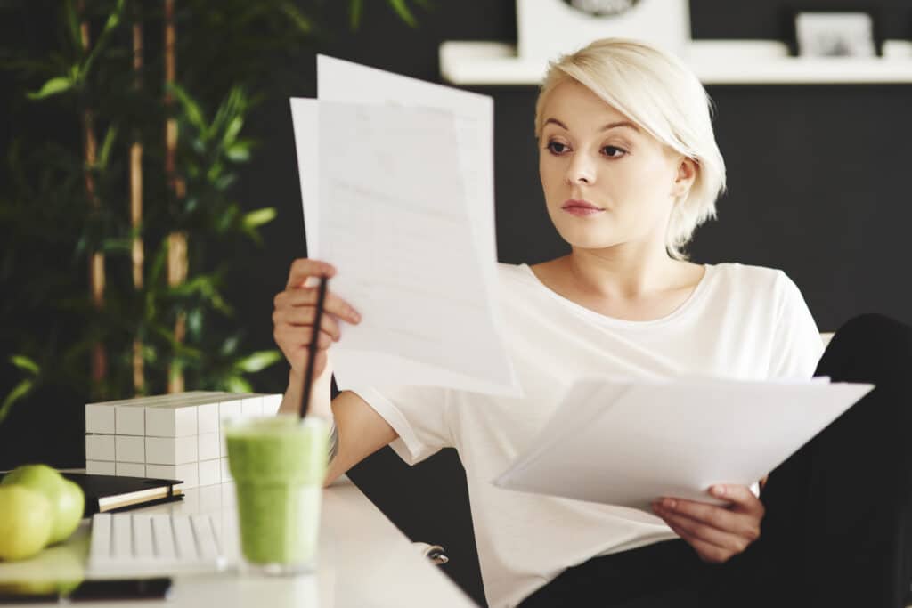 A blonde woman dressed up is reading documents while having a smoothie.