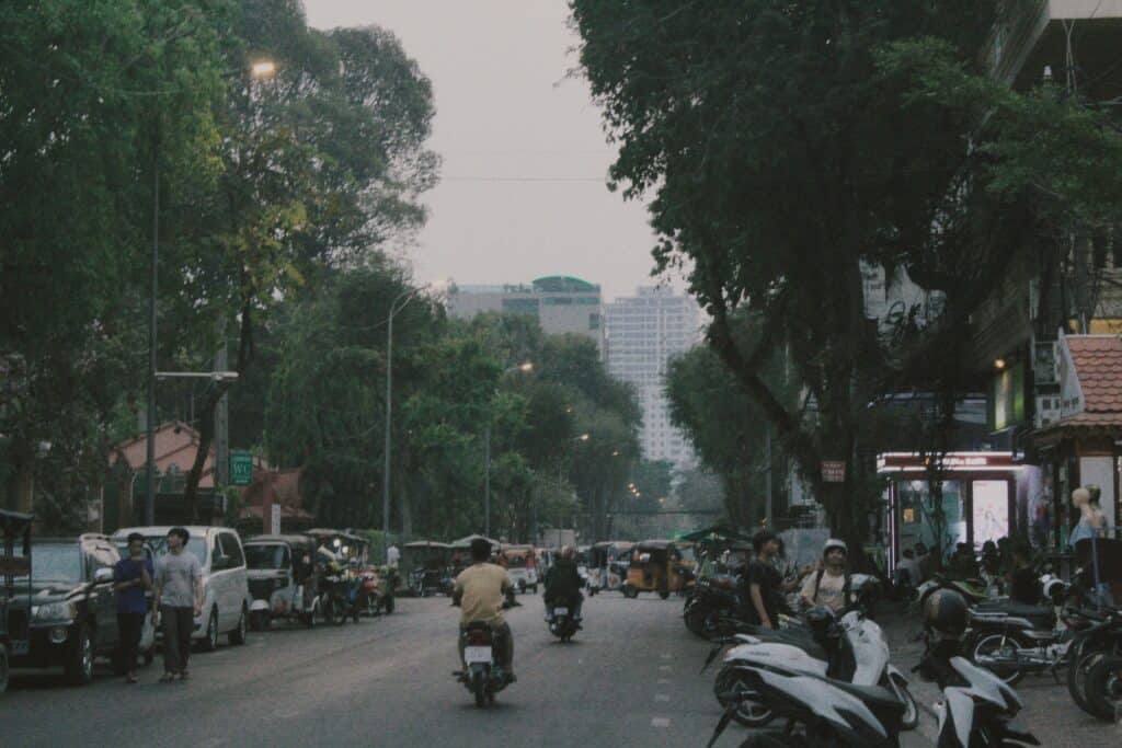 Crowded and busy street of Phnom Penh in Cambodia, with motorbikes on the road with cars parked alongside, and people chatting along the pavements.