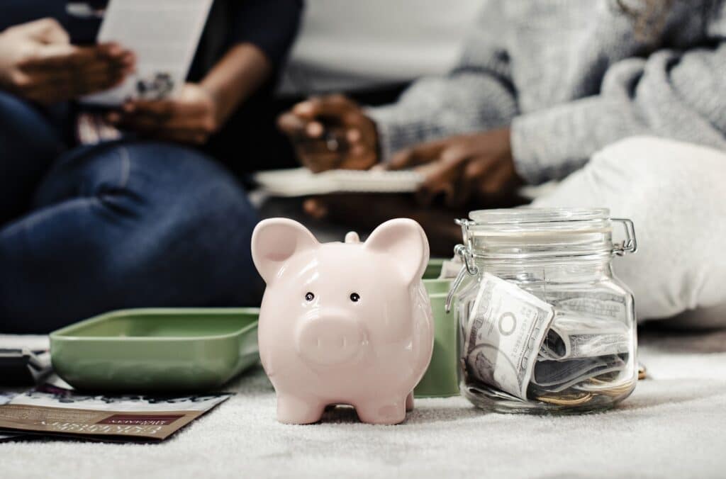 In the foreground is a piggy bank next to a jar filled with money, with the people sitting behind counting their finances to save money.