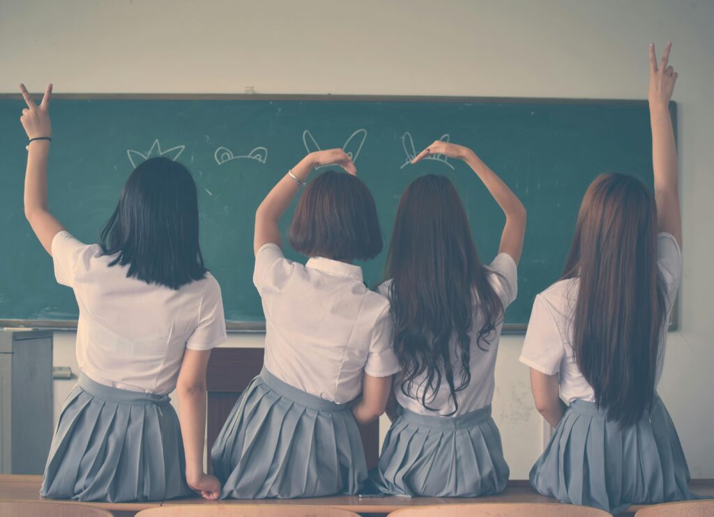 Four young Japanese schoolgirls in uniform sit on a classroom table, turn their backs and make heartfelt signs of affection with their hands and arms.