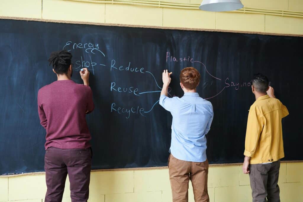 Three male students with their backs turned to a blackboard in a classroom, writing texts.