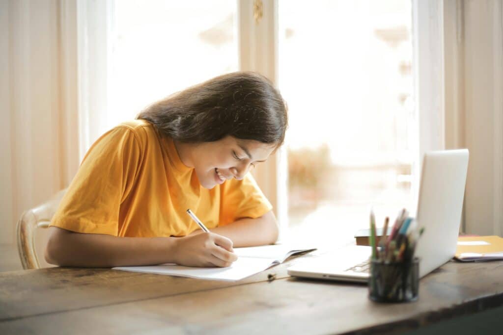 Young girl dressed in yellow happily doing her schoolwork. She is writing on a sheet of paper.