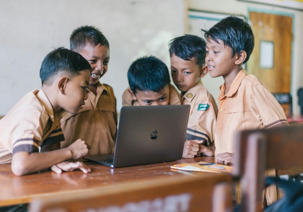 Young boys of South Asian origin in a classroom take an interest in a Macbook computer and look at what's on the screen with curiosity and interest.