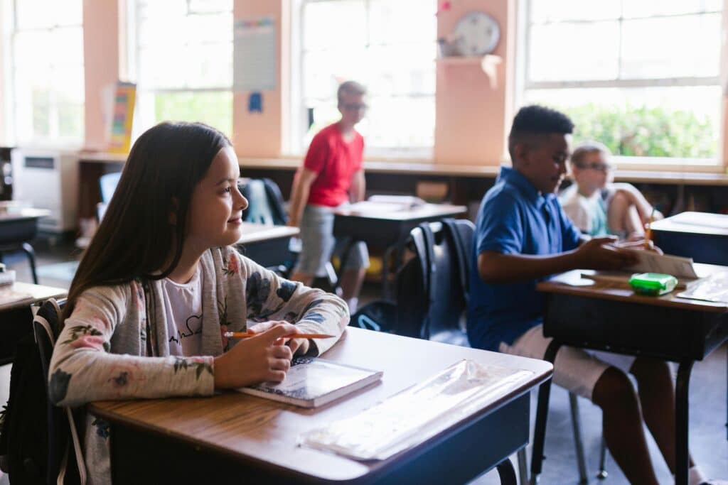 A small group of pupils in a classroom, seated at wooden desks. A young girl in the foreground is smiling and listening attentively to the teacher.