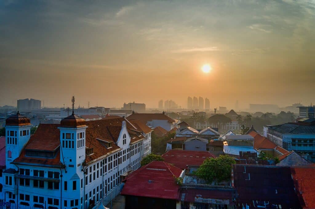 View from the topof a residential area in Jakarta. The street is paved and the surrounding houses are rustic, with red tiled roofs. There are some high buildings on the backgroud. The photo was taken at sunset, creating a peaceful atmosphere.