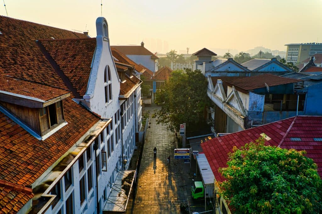 View of a residential street in Jakarta. The street is paved and the surrounding houses are rustic, with red tiled roofs. The photo was taken at sunset, creating a peaceful atmosphere.