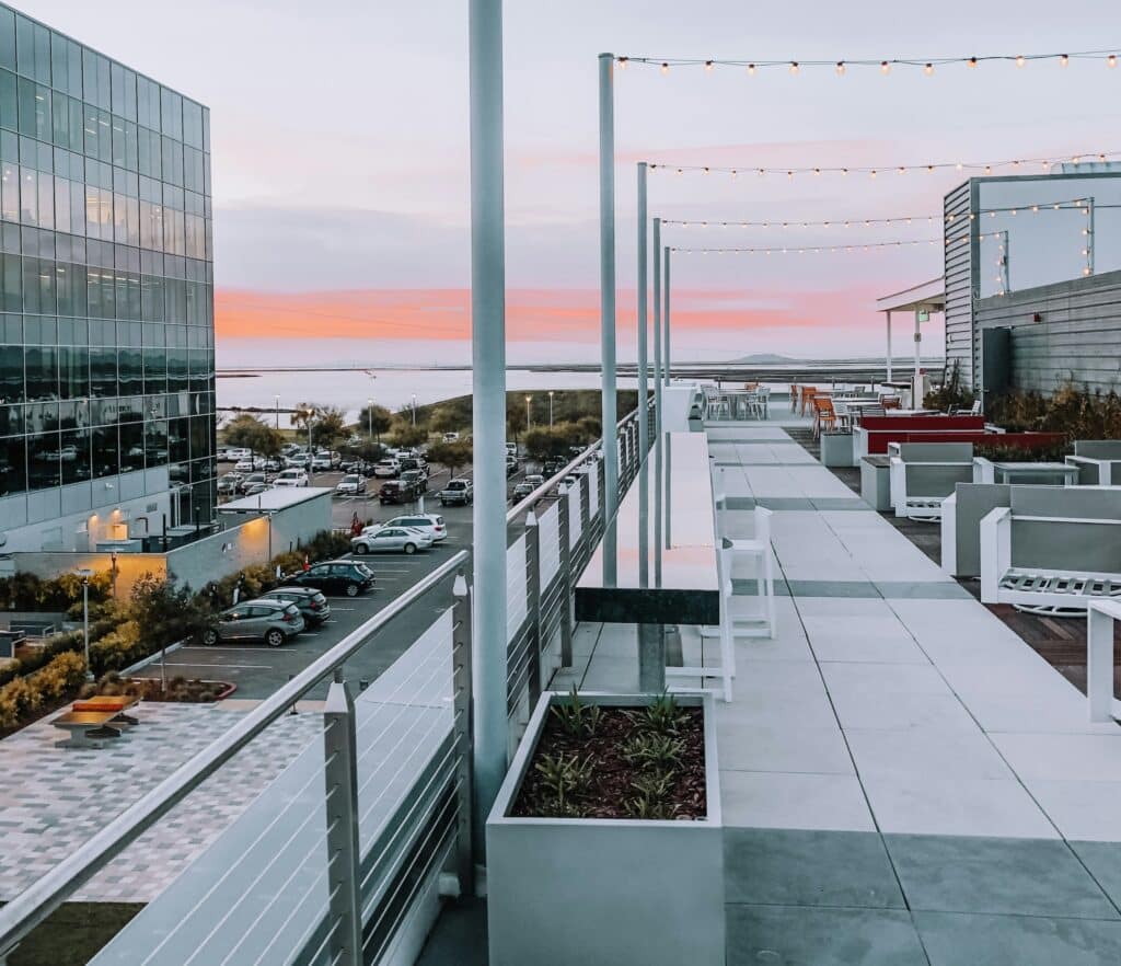 View of the roof terrace of a glass building in Silicon Valley in the United States. There is a car park at the foot of the building. The sky is a sunset shade of orange.