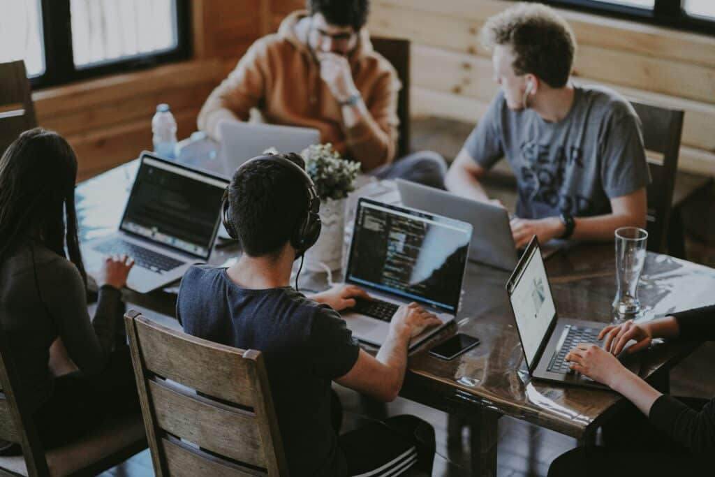 A group of young people working on their laptops around a table in a wooden chalet. Some are wearing headphones and chatting to each other.