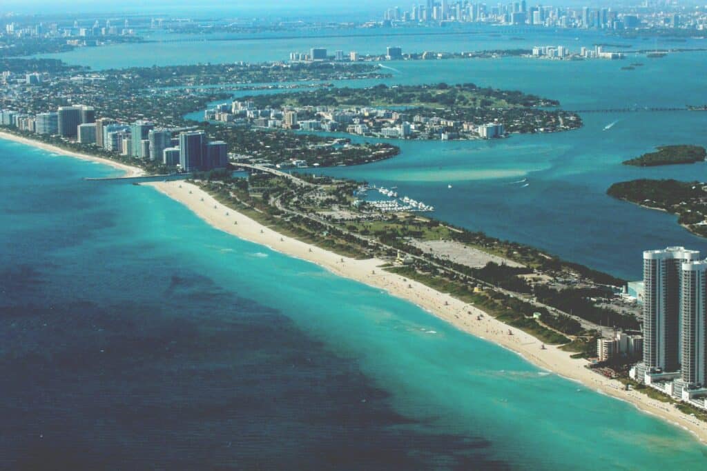 Aerial view of Miami in the United States. The turquoise-blue sea borders both sides of the mainland, where you'll find green spaces, tall buildings and a marina. The surrounding city can be seen in the background.