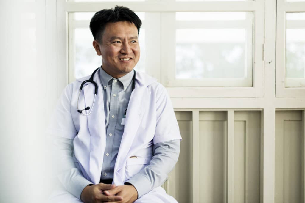 An Asian doctor sits smiling on a hospital windowsill. He has a stethoscope around his neck and is wearing a doctor's coat.