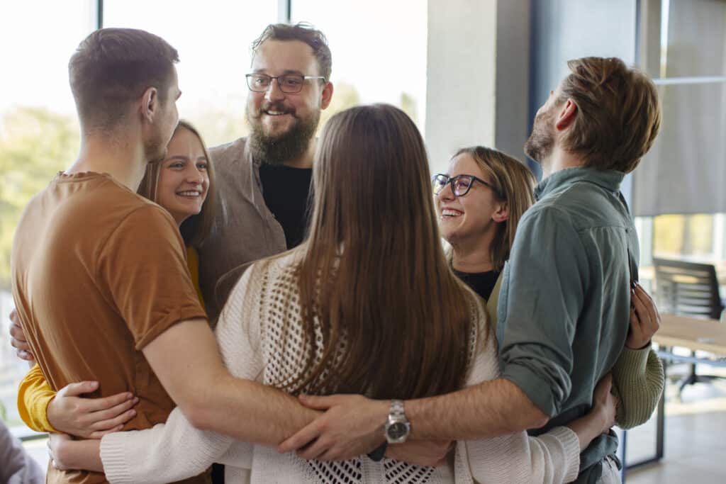 A group of young people form a circle and hold each other by the arms, smiling at each other in the spirit of caring.