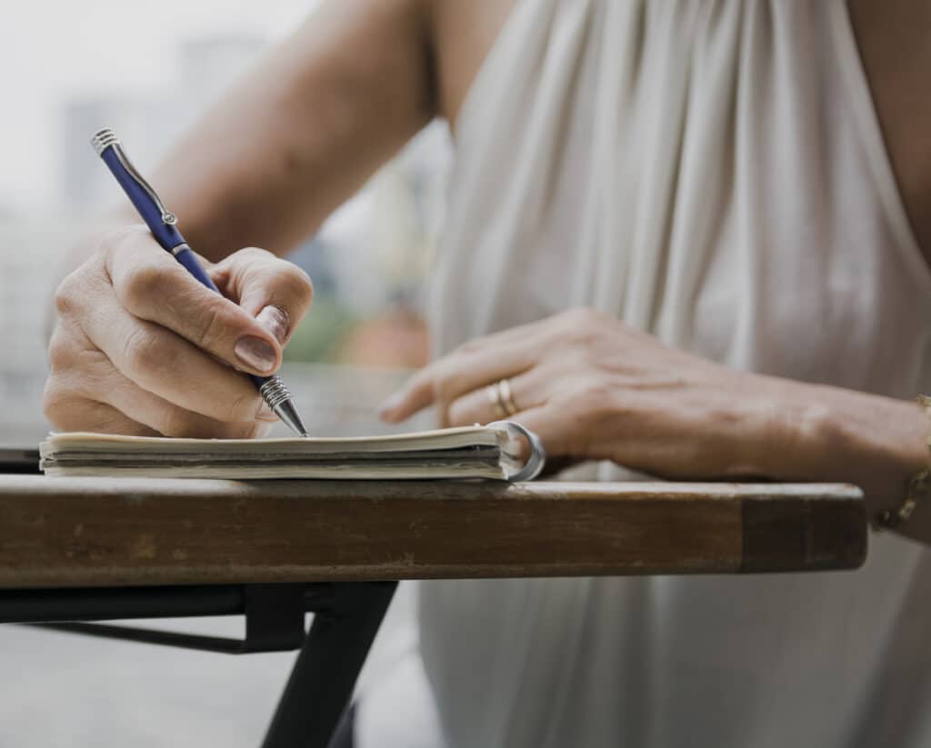 The photo shows a woman's hand taking notes in a notebook with a pen. The young woman is wearing a white sleeveless top and is sitting at a wooden table.