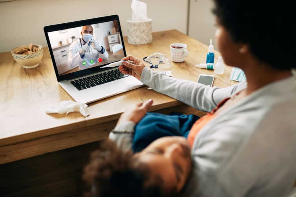 A mother of African origin sits on a chair and holds her daughter while she has a teleconsultation with a paediatrician. She holds a thermometer after taking her daughter's temperature.