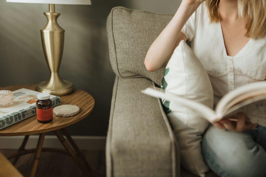 A woman in a white T-shirt is sitting on a sofa, reading a book and leaning back on a cushion. Next to the sofa is a side table containing a book, a lamp and a jar of food supplements.