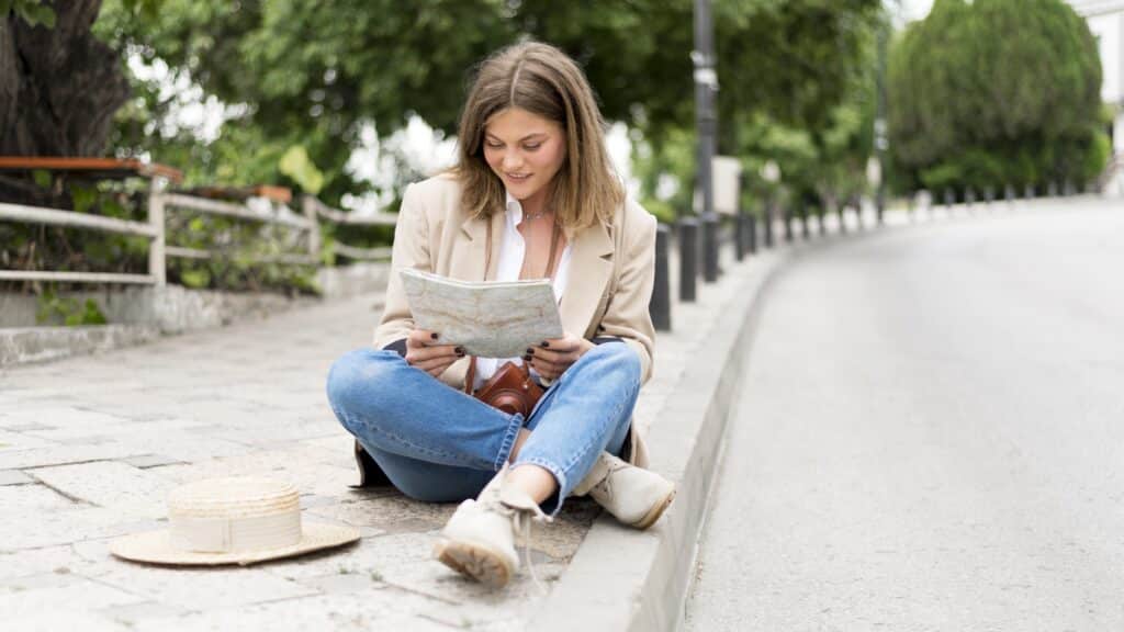 A young blonde woman is sitting on the pavement in the street, consulting a map of the city she is visiting. She has placed her hat on the ground.