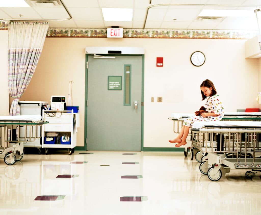 A little girl with a short bob sits on her hospital bed. She is alone in her hospital room, with its basic equipment and style.