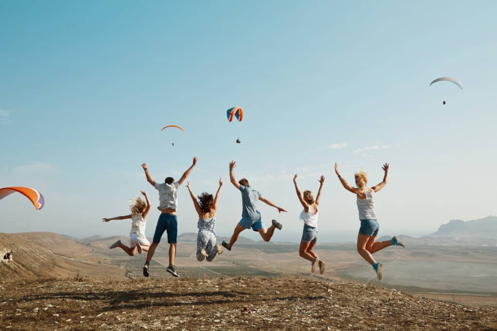 A group of friends are happily jumping into the air in a mountain setting. You can see the people from behind, and you can also see other people paragliding in the surrounding landscape. The sky is clear blue.