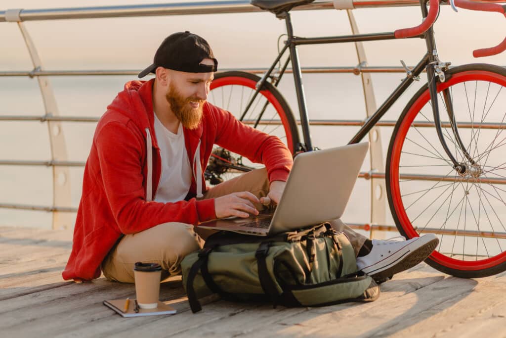 A red-haired man wearing a black cap and a red sweatshirt is typing on his laptop on top of his bag. The man is sitting cross-legged on the ground on a wooden bridge. His bike is lying next to him, along with a notebook and a takeaway coffee. The sea can be seen in the background and the scene was photographed during a level of sunshine.