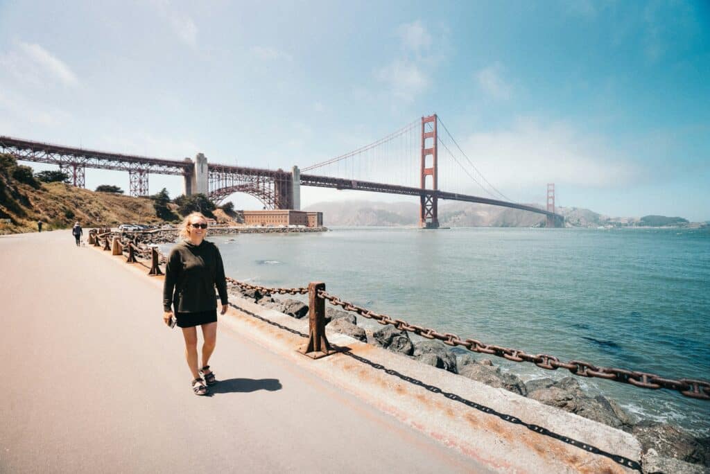 A woman in a black tracksuit walks along the seawall near the San Francisco bridge in the United States. She smiles at the caméera. The weather is splendid.