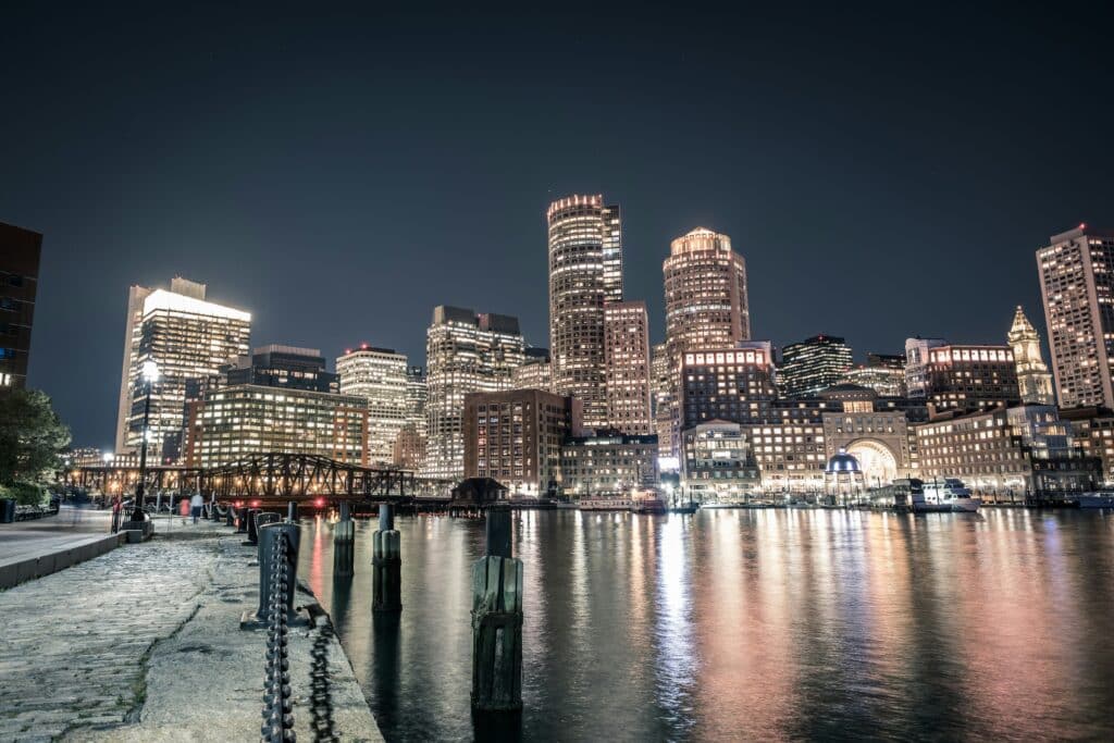 Night shot of the city of Boston in the United States and its river, where a few people are strolling along the banks. The buildings are illuminated, reflecting the atmosphere of a bustling, modern city.