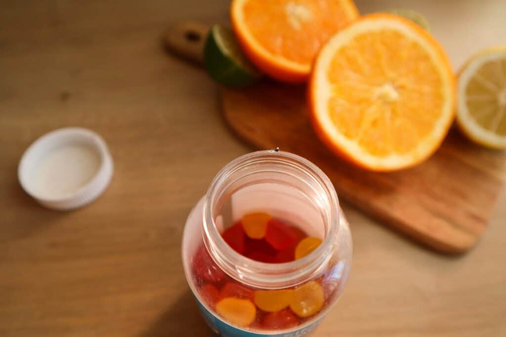 The photo is taken above an open jar of vitamins in the form of yellow and red gummies. Orange slices are displayed next to the jar.