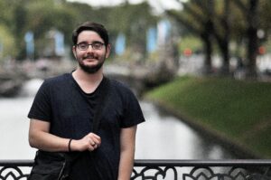 A bespectacled man in a T-shirt carries a bag and smiles at the camera. He is standing against the railing of a bridge.