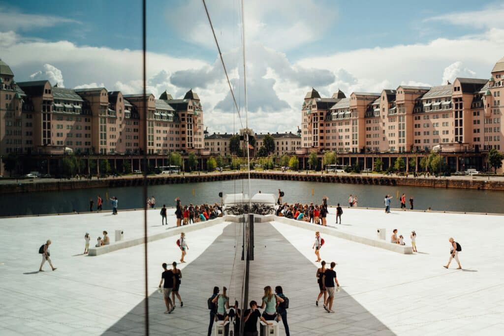 The photo was taken against a window in the Oslo Opera House in Norway to create a mirror effect. People are gathered in front of the opera house on the esplanade. There is a canal around the opera house surrounded by salmon-coloured buildings.