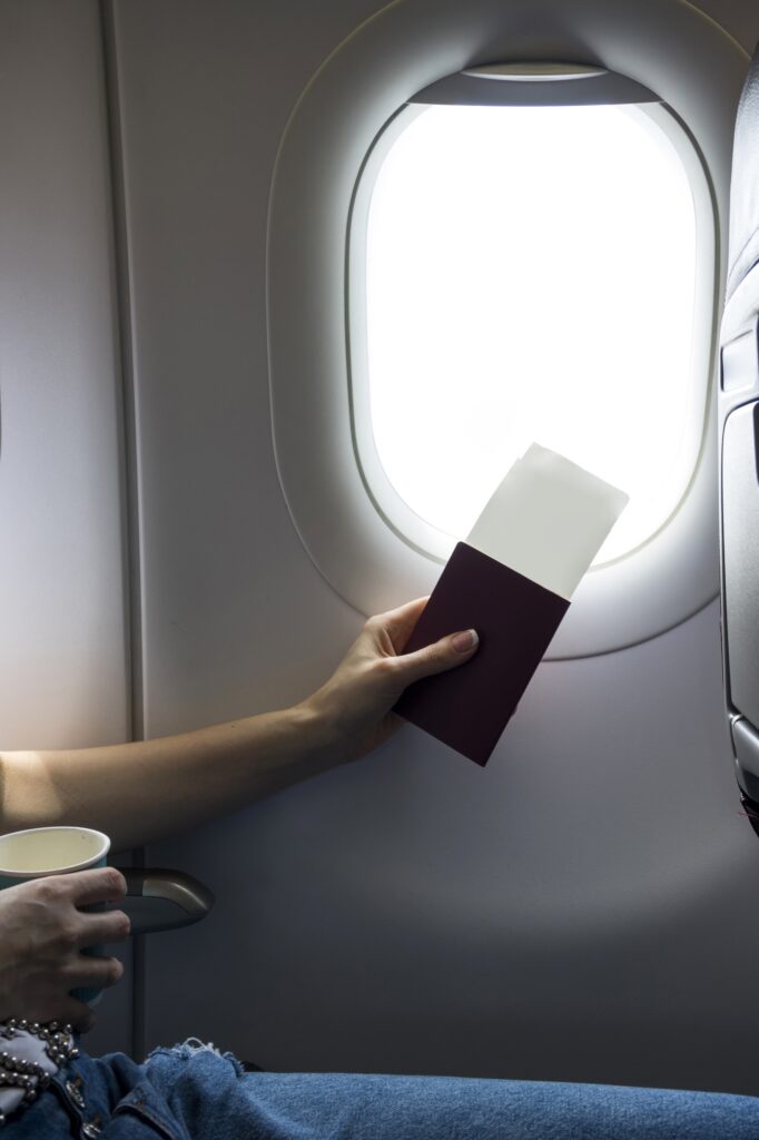 Hand holding a passport and plane tickets in front of the window of a plane in flight.