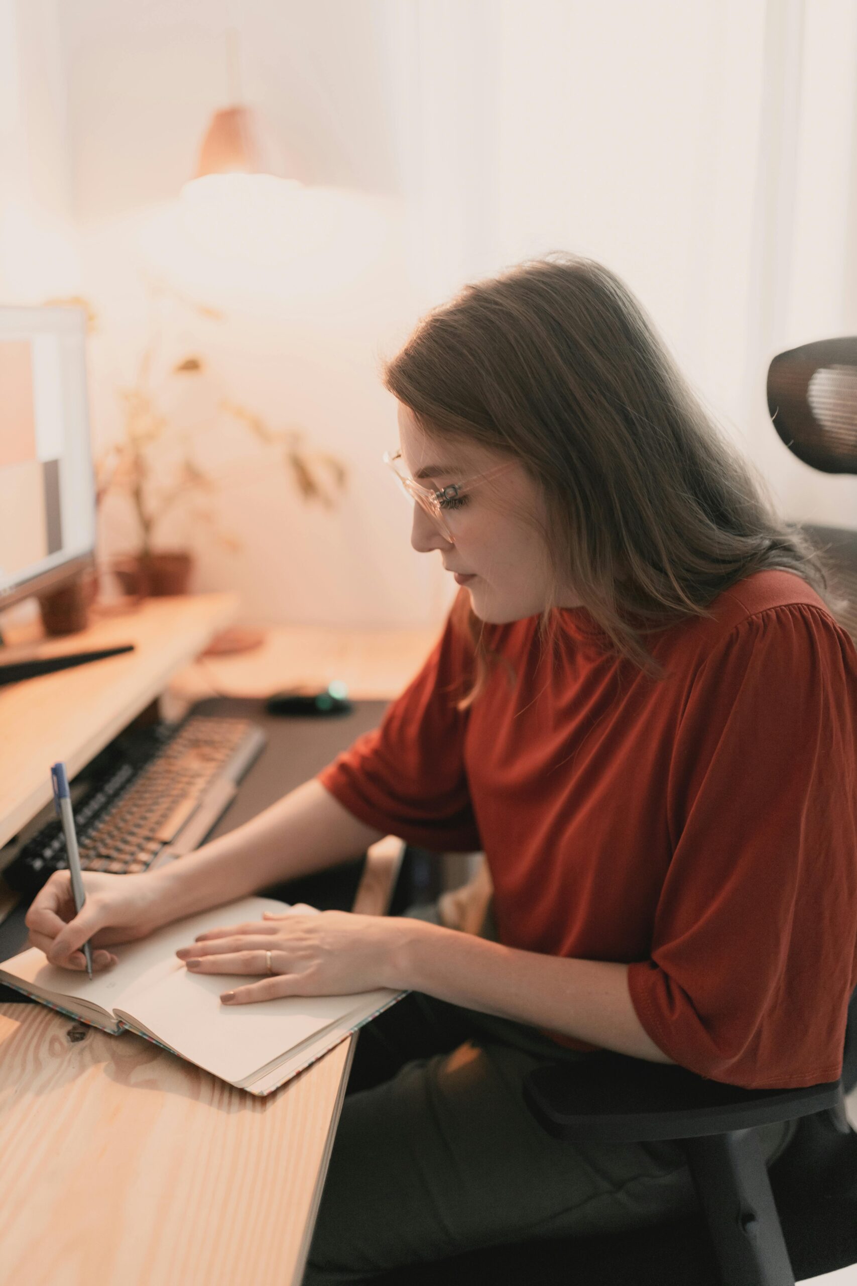 A young woman wearing glasses and a red blouse is concentrating on making notes in her notebook as she sits at her desk at home.