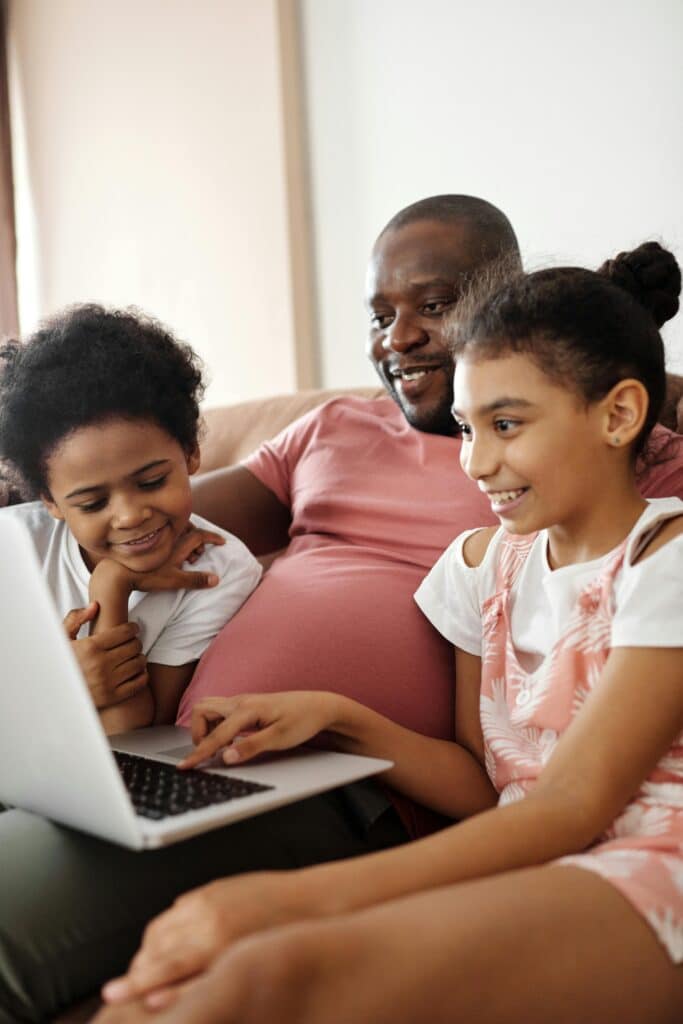 A father of African origin is sitting with his two daughters on the sofa in the living room, chatting to someone from a laptop computer on his lap. Everyone seems happy and in good spirits.