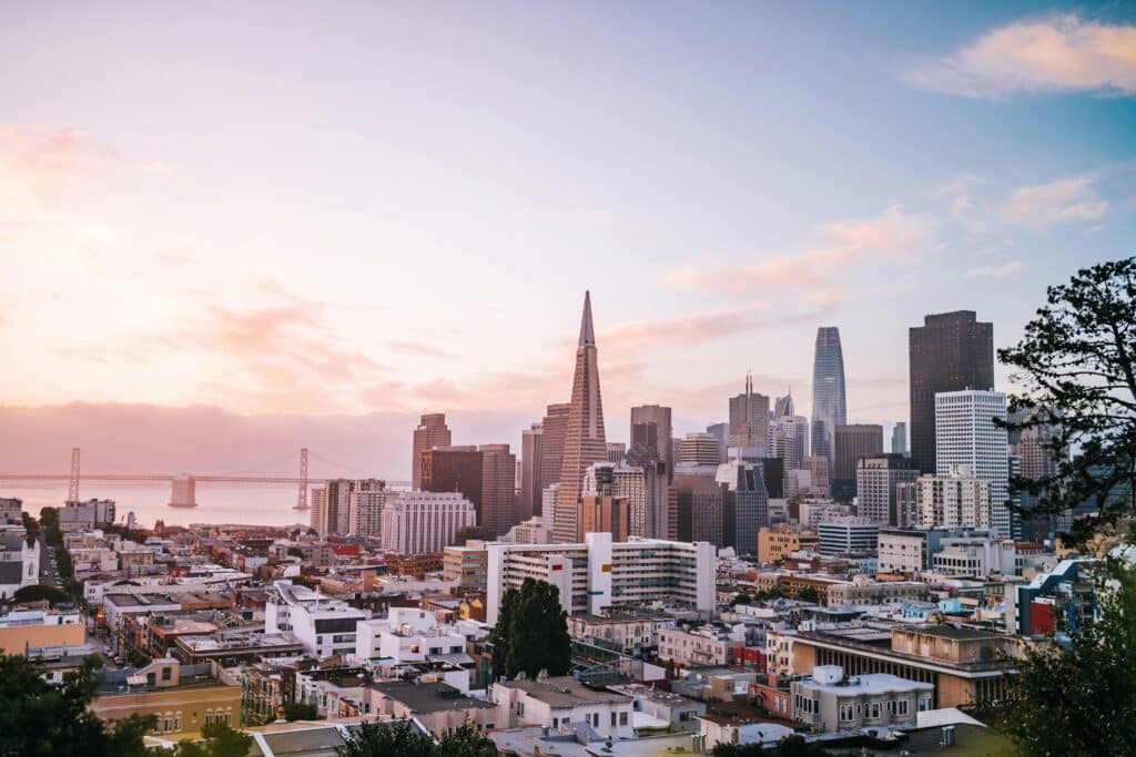 The beautiful city of San Francisco in the United States at sunset. The sky is painted in shades of pink and purple. In the foreground are small buildings, with skyscrapers in the background. The sea and the San Francisco Bridge can be seen in the background.