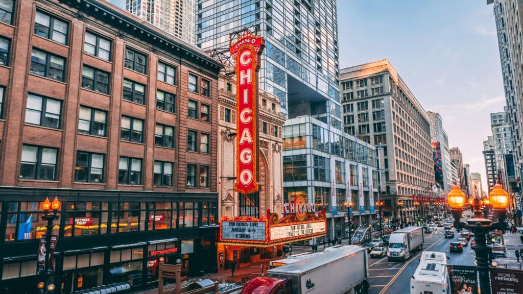 The photo shows one side of a street in Chicago in the United States. The street is packed with people and cars. The buildings are modern, and the theatre has a large illuminated sign forming the word Chicago. The headline show is Paw Patrol live. The photo was certainly taken at the end of the day, judging by the pinkish colour of the sky.