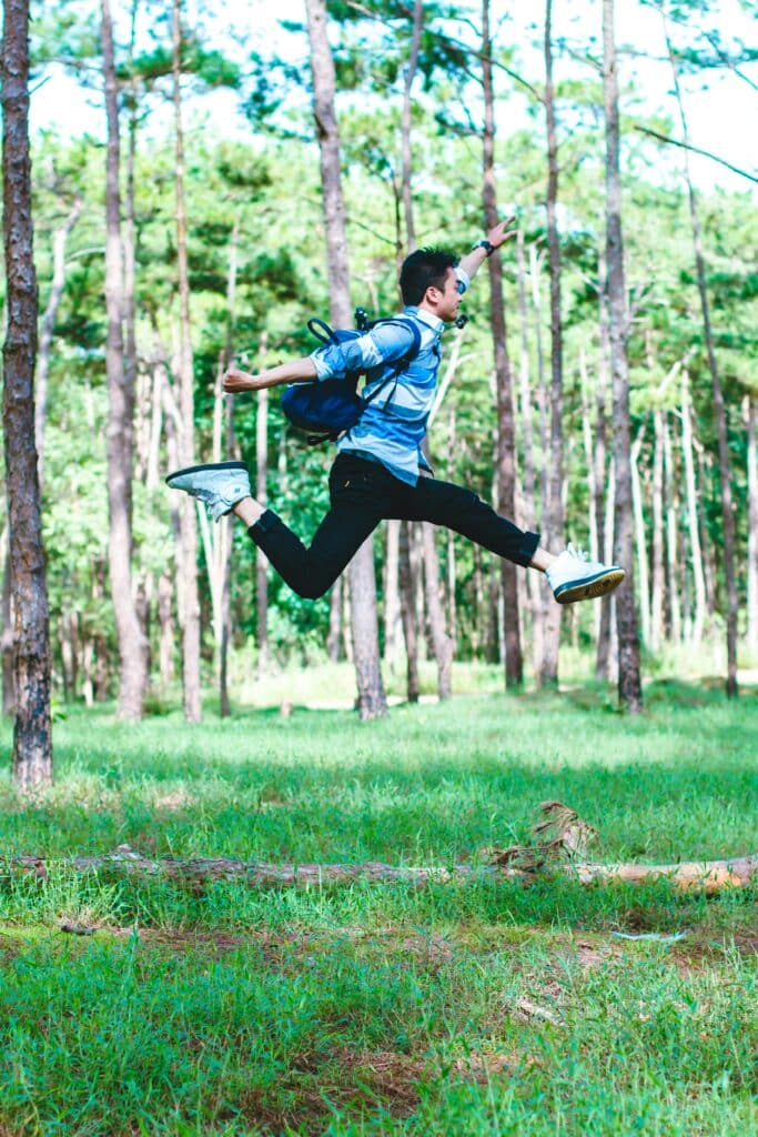 A young Asian man jumps into the air in the middle of a forest. He is wearing a blue shirt and rucksack, a pair of black jeans and white trainers. The weather is sunny.