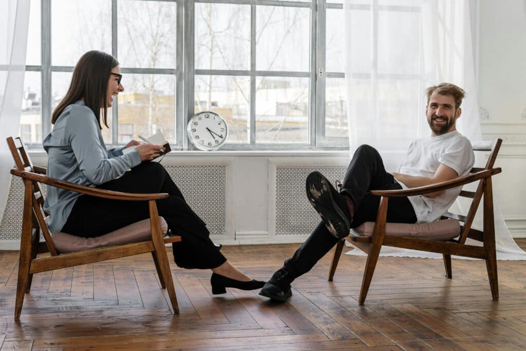 A smiling young man is in the middle of a psychotherapy session with his psychologist. They are both seated on modern wooden chairs in a room with white walls and dark wood parquet flooring. The young female psychologist is also laughing. She is holding a notebook and wearing a light blue shirt, black trousers and dark glasses. Her patient is wearing a white T-shirt and black trousers.