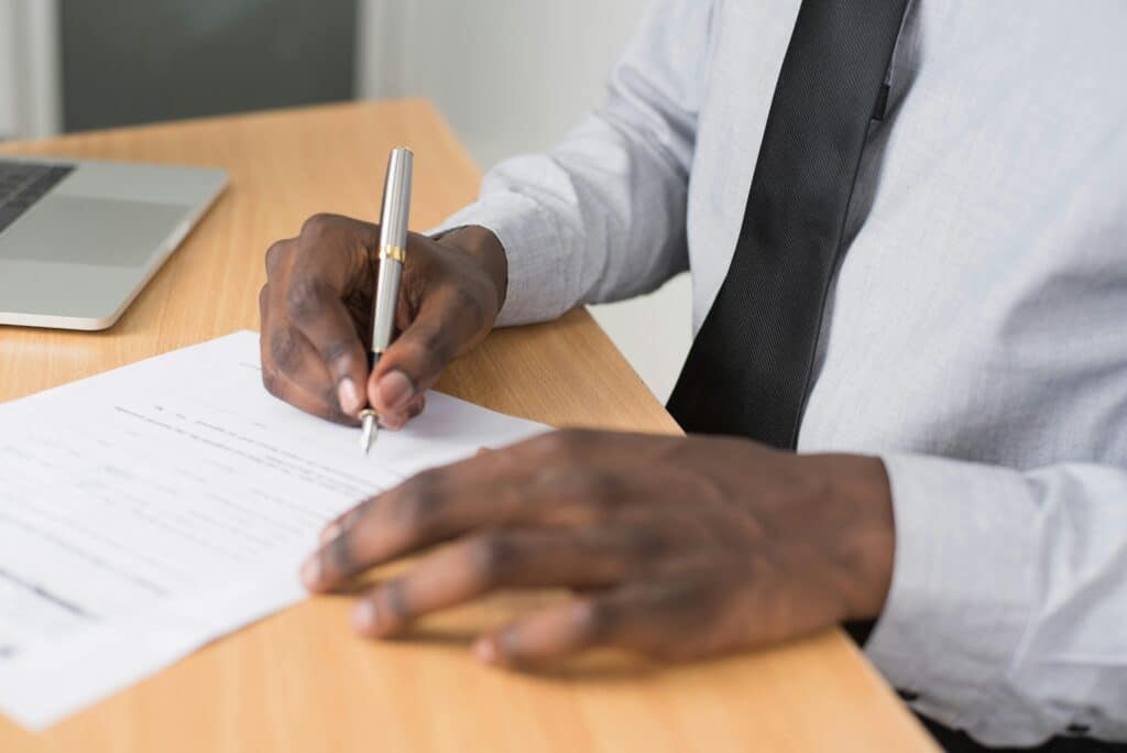 A black man in a suit and tie, whose face cannot be seen, only his hands, signs a document with a silver pen. A laptop sits beside him on his desk.