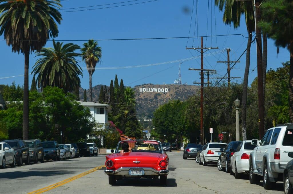 A young woman is driving a luxurious red car on a road in Los Angeles, USA, on a very sunny day. The famous Hollywood sign can be seen in the background of the photo, on the hillside.