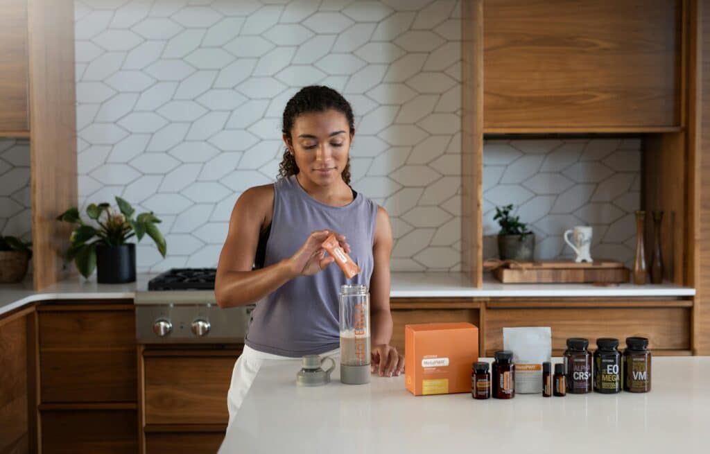 A young woman of African origin prepares a dietary supplement in the form of a powdered solution in a flask. Wearing a sports outfit, she mixes the mixture on the island in her modern wood and marble kitchen.