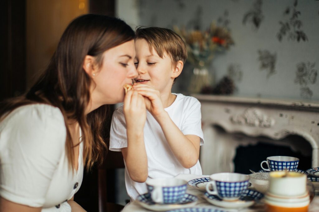 A young boy, sitting at table with his mother, feeds her with both hands. The table and dining room are decorated in a rustic, country style.