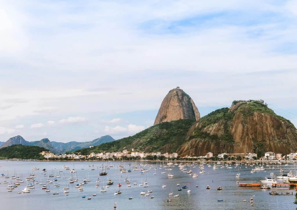 View of Sugarloaf Mountain and the beach in the Urca district of Rio de Janeiro, under a clear sky. The calm sea is full of small boats.