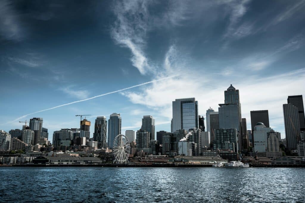 Photo of the port and city of Seattle in the United States, taken at sea level. The weather looks rough and a plane has just crossed the sky. Tall buildings dominate the landscape. There is a Ferris wheel along the coast.