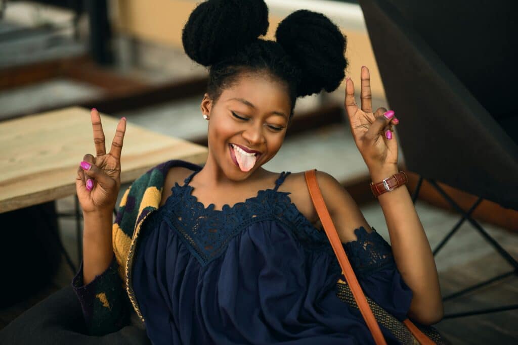 A woman smiles as she sticks out her tongue and closes her eyes. She is wearing a navy blue blouse and is making a victory sign with both hands.