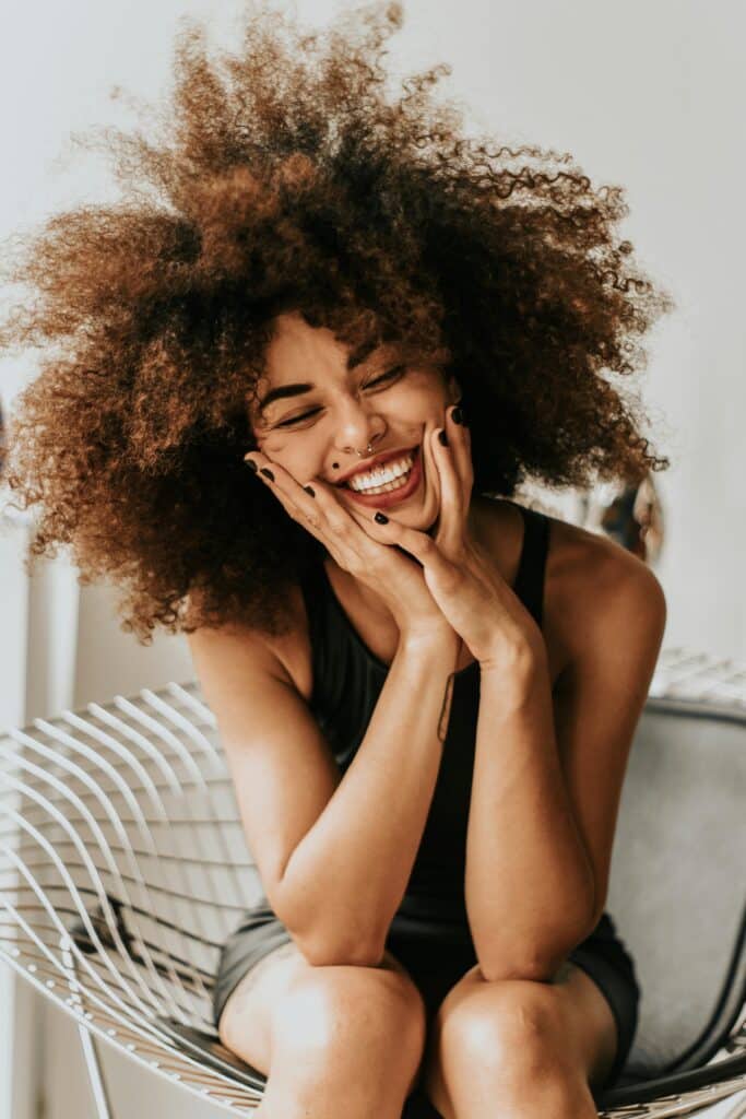 A beautiful young woman with an afro ocupe is smiling broadly, holding her face with her hands as she sits on a white metal chair. She has tattoos and a nose piercing. She is wearing a simple black tank top.